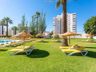 Grüner Rasen mit Liegestühlen und Strohhüte unter sonnigem Himmel, im Hintergrund Hochhäuser.
