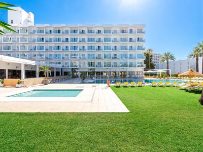 Hotelgebäude mit weißen Balkonen, großem Pool und grüner Liegewiese unter klarem Himmel.