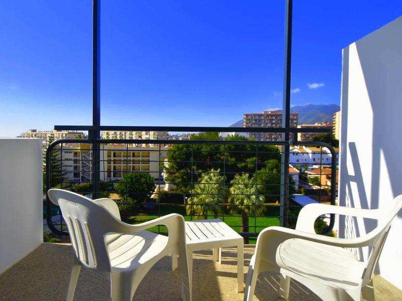 Balkon mit weißen Kunststoffstühlen und kleinem Tisch, dahinter Grünfläche und Gebäude, blauer Himmel.
