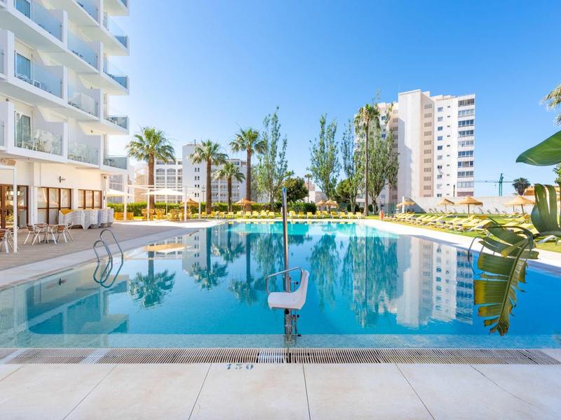 Moderner Außenpool mit klar blauem Wasser, umgeben von weißen Gebäuden und Palmen unter blauem Himmel.