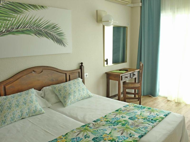 Helles Schlafzimmer mit zwei Betten, Holzkopfteil, floralem Bettläufer und grünem Wanddekor.