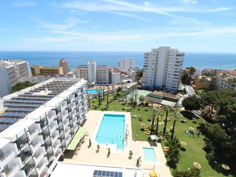 Blick auf Hotel mit Pool, grüner Gartenanlage und Hochhäusern nahe dem Meer und blauem Himmel.