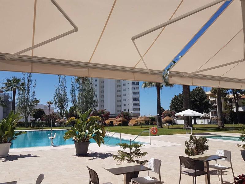 Moderner Außenpool mit Liegestühlen, Pflanzen und Sonnenschirm vor hohem Wohnhaus bei klarem Himmel.