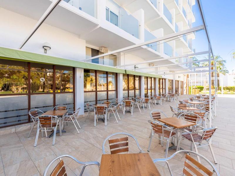 Freiluftterrasse mit mehreren Holztischen und Metallstühlen vor modernem weißen Gebäude.