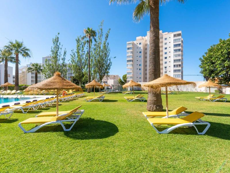 Grüner Rasen mit Liegestühlen und Strohhüte unter sonnigem Himmel, im Hintergrund Hochhäuser.