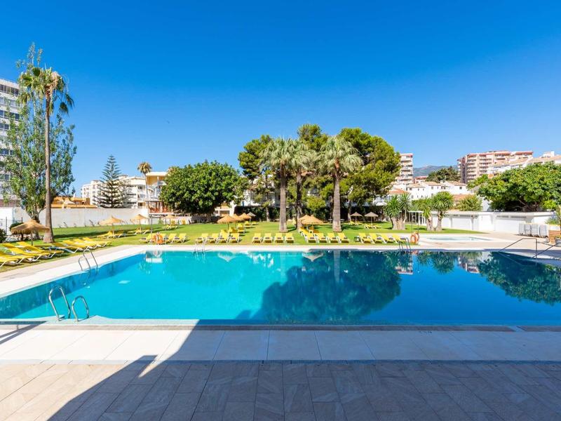 Freier Swimmingpool mit klarem Wasser, umgeben von Liegestühlen und Palmen bei blauem Himmel.