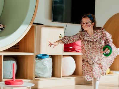 Kind wirft Ringe in farbige Stäbe in einem hellen Raum mit Holzregalen und buntem Spielzeug.