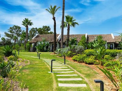 Ein Gartenweg mit rechteckigen Steinen führt durch grüne Pflanzen zu einem Haus mit Palmen.
