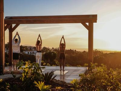 Tre persone fanno yoga su una terrazza con vista sul tramonto e un paesaggio rurale.