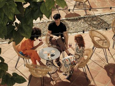 Drei Personen sitzen an einem runden Tisch mit Getränken auf einer sonnigen Terrasse mit Stühlen.