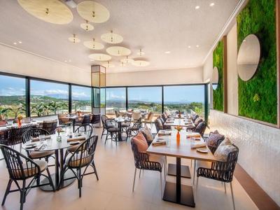 Helles modernes Restaurant mit großen Fenstern, Holz- und Metallstühlen, grüner Wanddekoration und weißen Tischen.