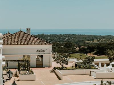 Blick auf eine Terrasse mit weißen Gebäuden, grünen Pflanzen und blauem Himmel am Meer im Hintergrund.