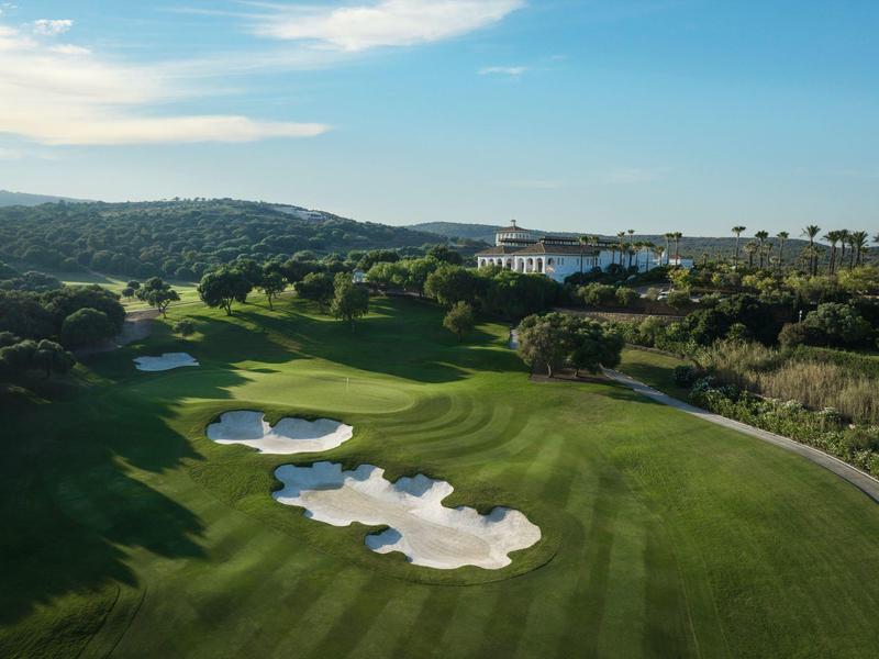Golfplatz mit mehreren Sandbunkern, grünen Fairways und bewaldetem Hintergrund unter blauem Himmel.