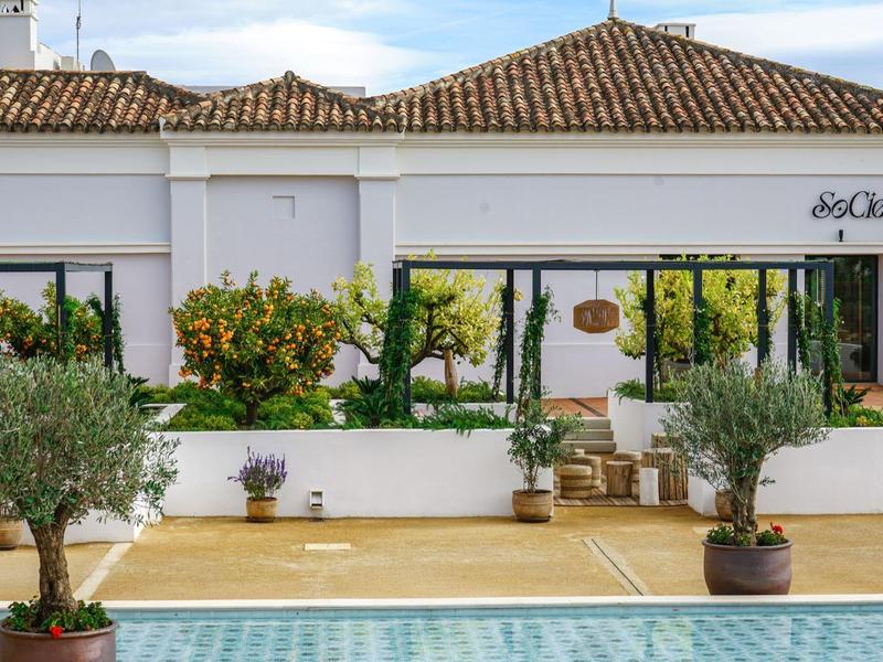 Weißes Gebäude mit braunem Ziegeldach, Blumen und Pflanzen vor einer verglasten Veranda.