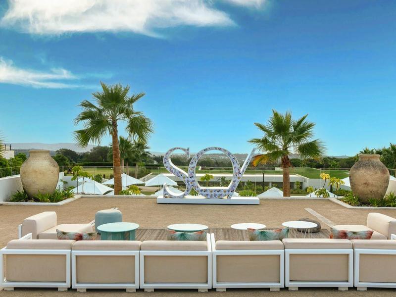 Moderner Außenbereich mit Sitzmöbeln, Palmen, dekorativem Schriftzug "sea" und blauem Himmel.