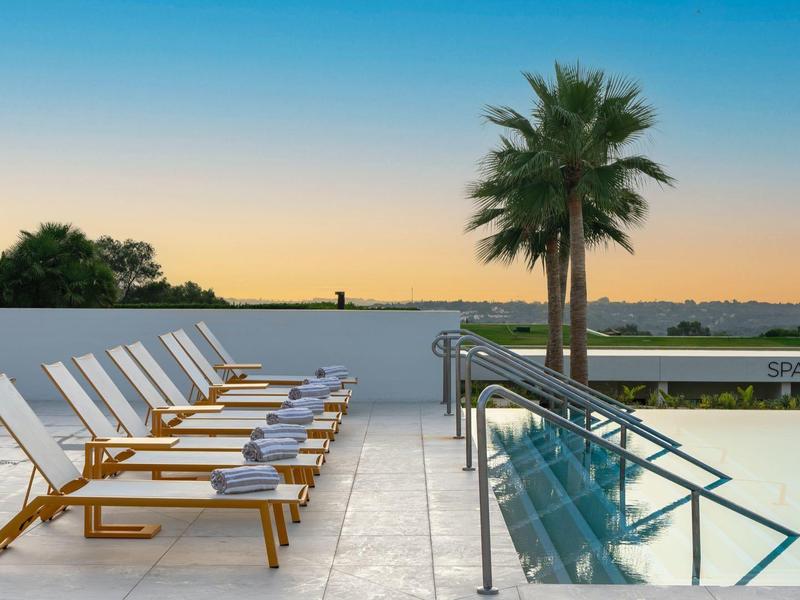 Pool mit glasklarem Wasser, Liegestühlen mit blauen Handtüchern, Palmen und Sonnenuntergang.