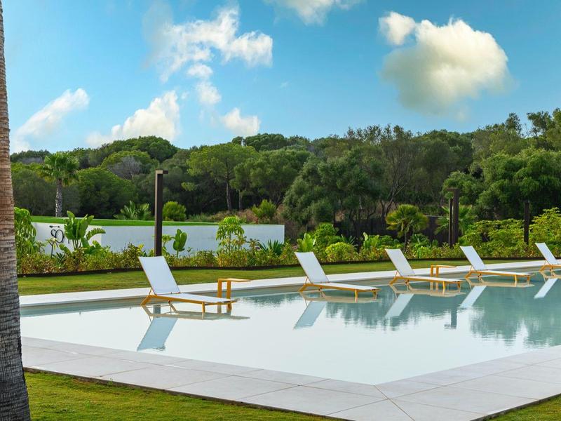 Moderner Pool mit weißen Liegestühlen, Palmen und grünem Hintergrund unter blauem Himmel.