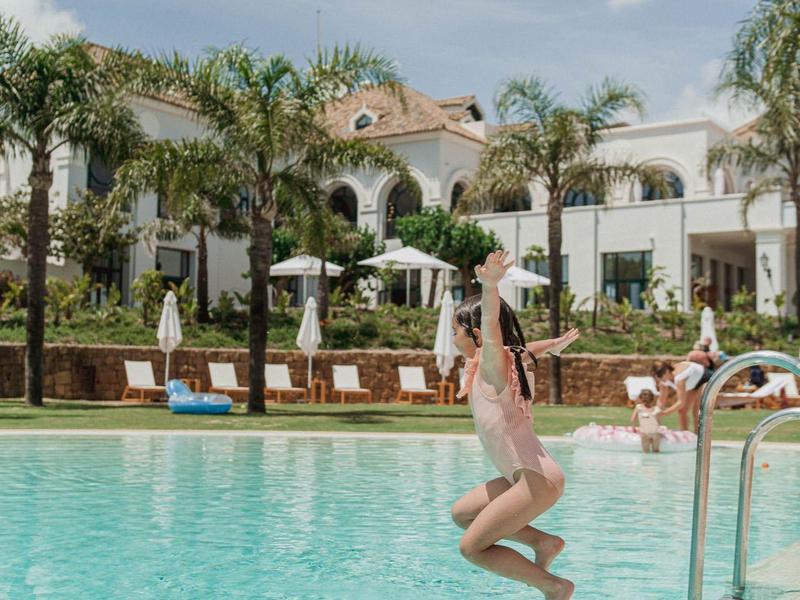 Ein Mädchen springt ins türkisfarbene Schwimmbad vor einem weißen Hotel mit Palmen und blauem Himmel.