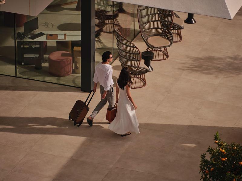 Zwei Personen mit Rollkoffern laufen über einen sonnigen, offenen Platz neben einem Glasgebäude.