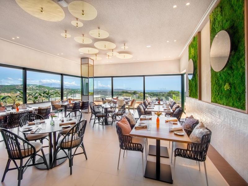 Helles modernes Restaurant mit großen Fenstern, Holz- und Metallstühlen, grüner Wanddekoration und weißen Tischen.