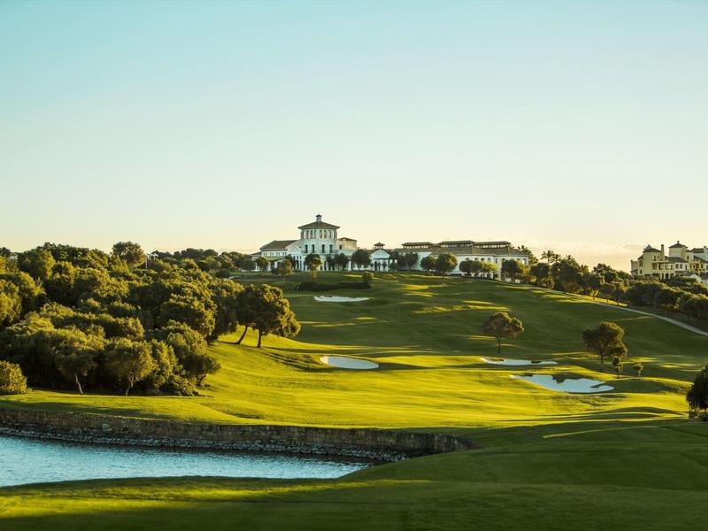 Sanfter Hügel mit grünem Rasen, Bäumen, Wasser und Gebäuden bei klarem Himmel am Abend.