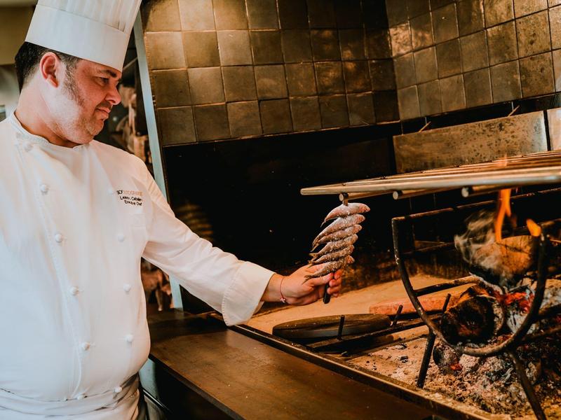 Koch in weißer Uniform hält einen Entenspieß vor einem offenen Grill mit brennenden Holzscheiten.