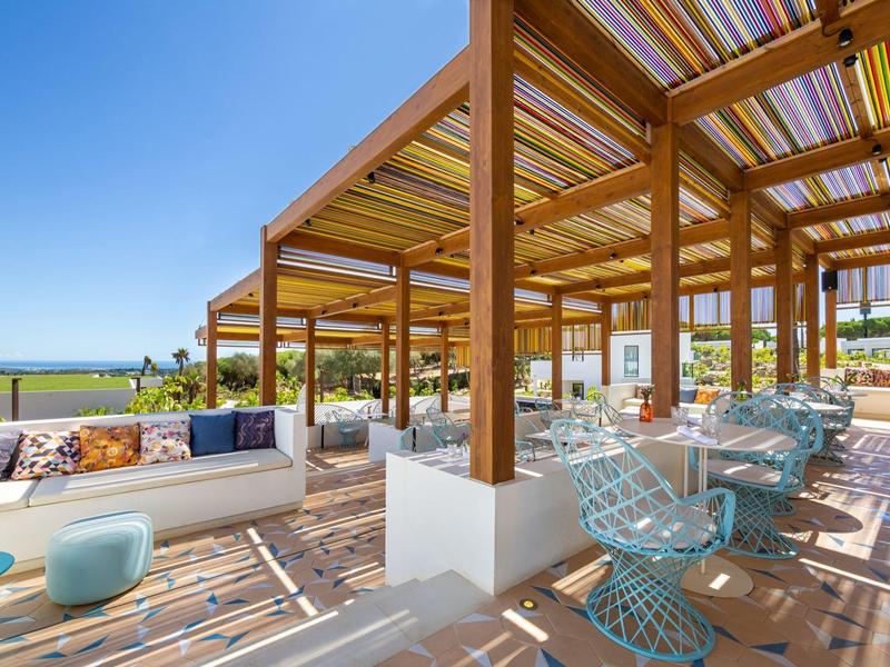 Offener Sitzbereich mit Holzstruktur, bunten Stühlen und Sofas bei klarem Himmel.