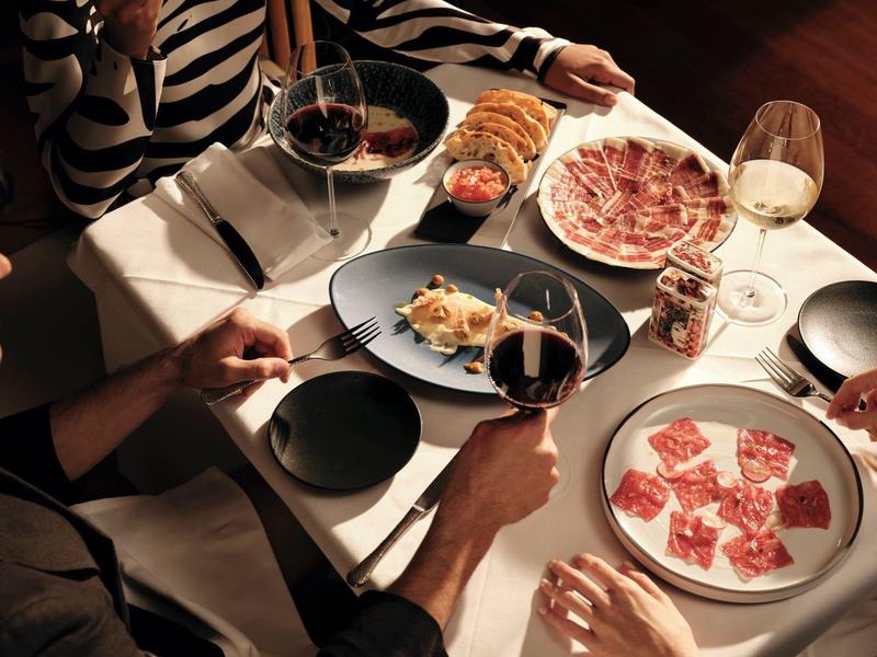 Drei Personen genießen gemeinsames Raclette-Essen an weiß gedecktem Tisch mit Wein.
