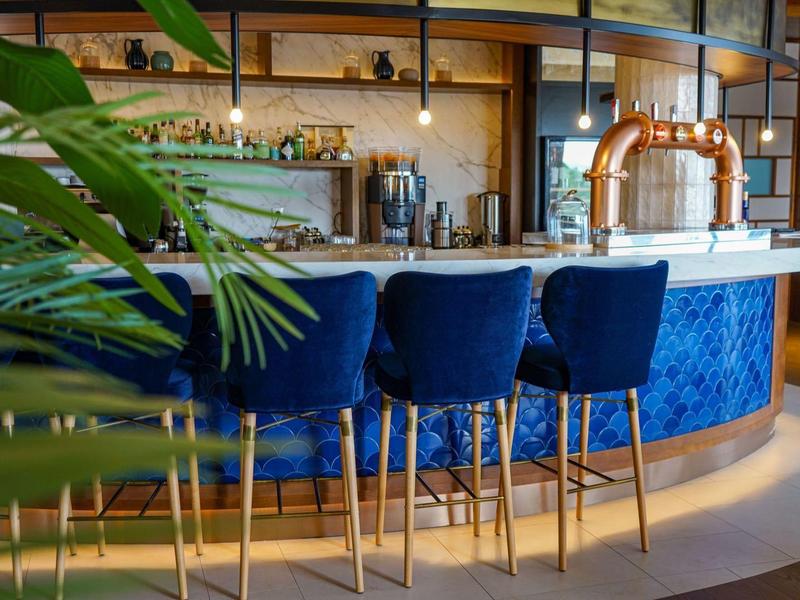 Moderne Bar mit blau gepolsterten Hockern, Kupfer-Akzenten und grüner Pflanze im Vordergrund.