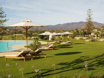 Grüne Rasenfläche mit weißen Sonnenschirmen, Liegestühlen und Pool vor Bergen im Hintergrund.
