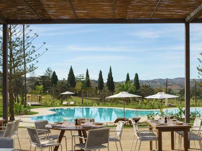 Terrasse mit Holzstühlen, Tischen, Pool, Sonnenschirmen, Blick auf grünes Feld und Bäume.