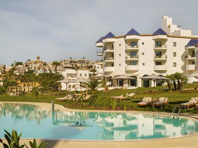 Großer blauer Pool vor weißem Hotelgebäude mit blauen Dächern und vielen Liegen im Garten.