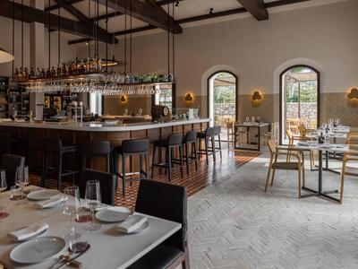 Moderne Restaurantbar mit Holztheke, hohen Stühlen, großen Fenstern und gedeckten Tischen im hellen Raum.