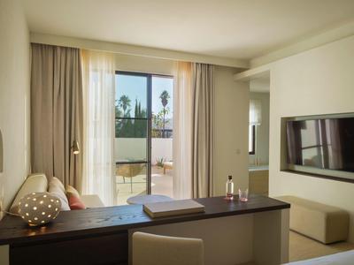 Helles Zimmer mit Schreibtisch, Fernseher an der Wand und Balkon mit Glasfenstern.