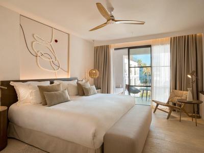 Helles Schlafzimmer mit Doppelbett, großen Fenstern, Gardinen und modernem Wandbild in neutralen Farbtönen.