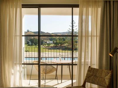Blick durch großes Fenster auf Balkon mit Stühlen und Tisch, dahinter See und bewaldeter Hügel.