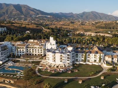 Großes weißes Hotel mit Pool, Grünflächen und Bergen im Hintergrund unter blauem Himmel.