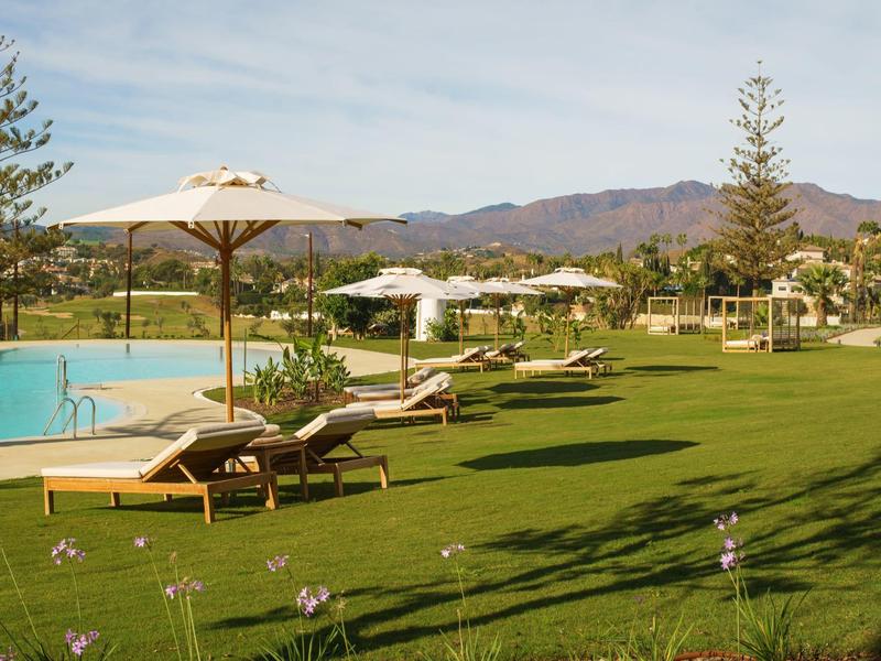 Grüne Rasenfläche mit weißen Sonnenschirmen, Liegestühlen und Pool vor Bergen im Hintergrund.