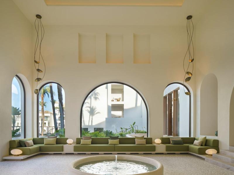 Großer Raum mit runden Fenstern, langem grünen Sofa und rundem Tisch in der Mitte unter hohen Decken.