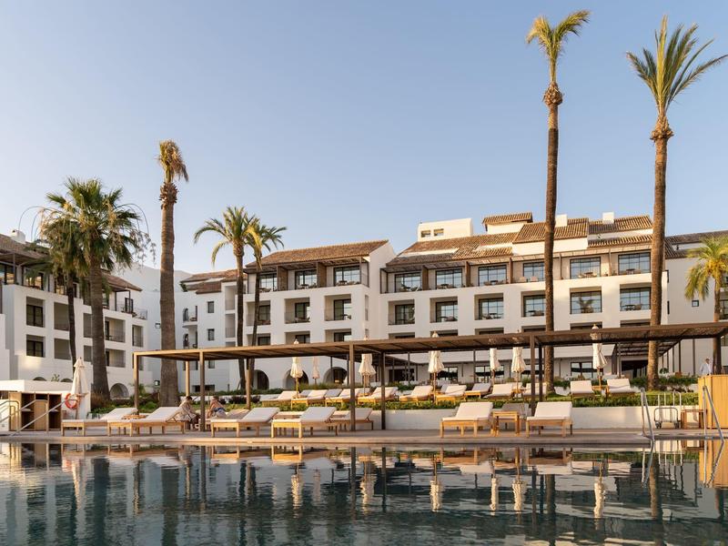 Ein modernes Hotel mit mehreren Stockwerken, Palmen und einem großen Pool im Vordergrund.