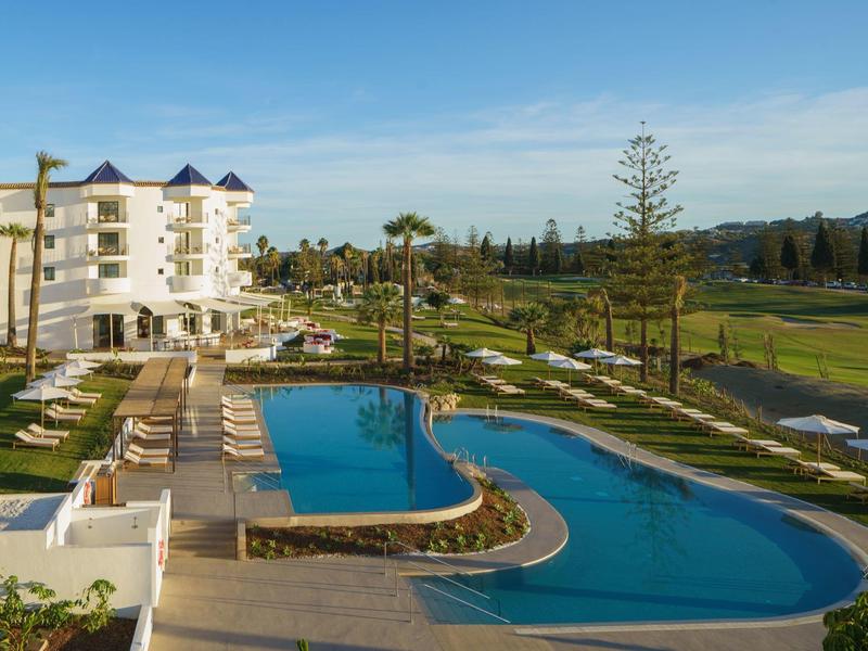 Schwimmbad mit geschwungener Form, umgeben von Liegestühlen und Palmen vor weißen Gebäuden.