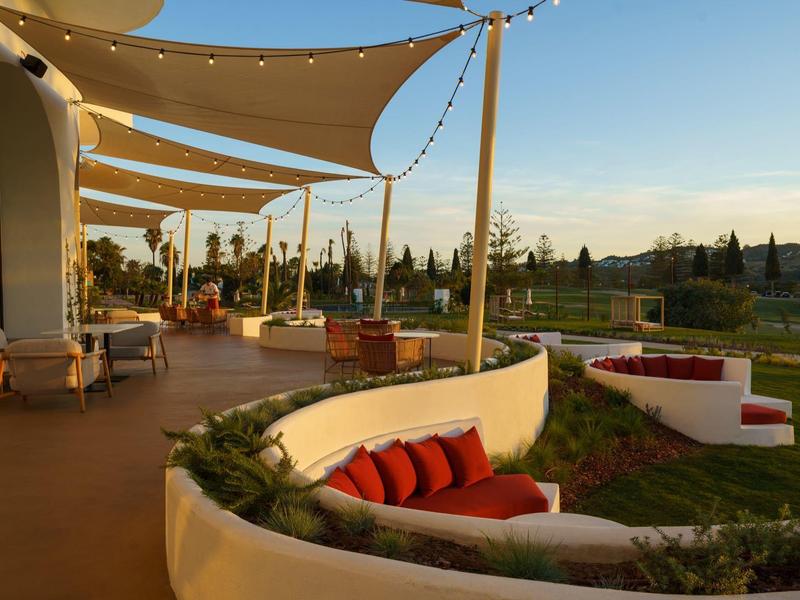 Terrassenbereich mit geschwungenen Sitznischen, roten Kissen und Sonnensegeln bei Sonnenuntergang.