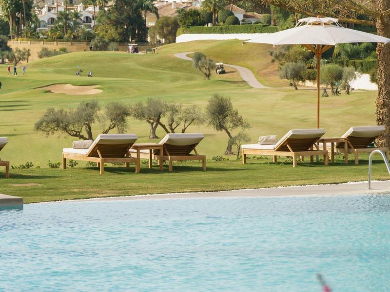 Schwimmbecken mit blauer Wasserfläche, Liegestühlen auf grünem Rasen und Sonnenschirm im Hintergrund.