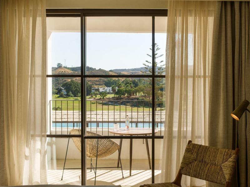 Blick durch großes Fenster auf Balkon mit Stühlen und Tisch, dahinter See und bewaldeter Hügel.