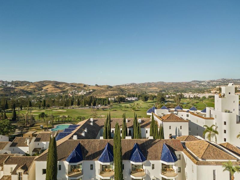 Stadt mit weißen Gebäuden, blauen Dächern, grünen Bäumen und Hügeln unter klarem blauem Himmel.