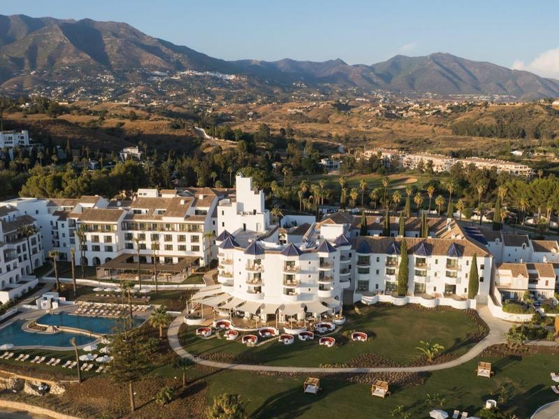 Großes weißes Hotel mit Pool, Grünflächen und Bergen im Hintergrund unter blauem Himmel.