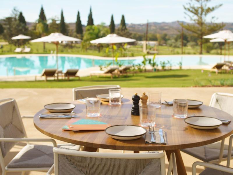 Runder Holztisch mit sechs gedeckten Plätzen im Freien, Hintergrund mit Pool und Sonnenschirmen.