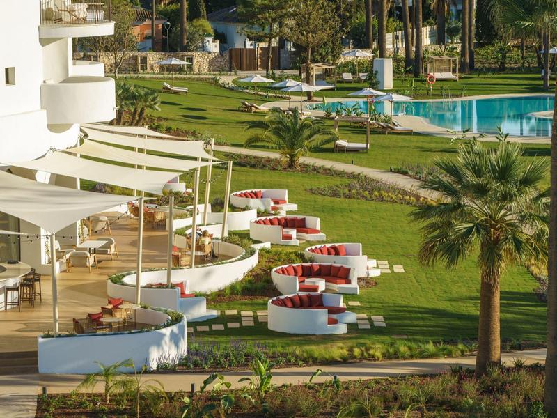 Grüne Gartenanlage mit weißen Loungemöbeln und Pool neben weißem Hotelgebäude bei Sonnenschein.