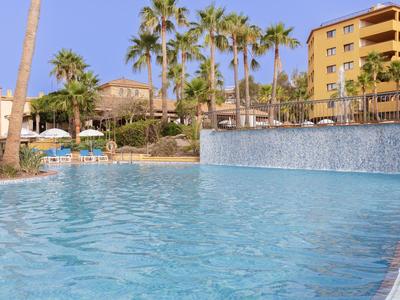 Großer Pool mit klarem Wasser, Palmen und gelben Gebäuden im sonnigen Resort-Hintergrund.