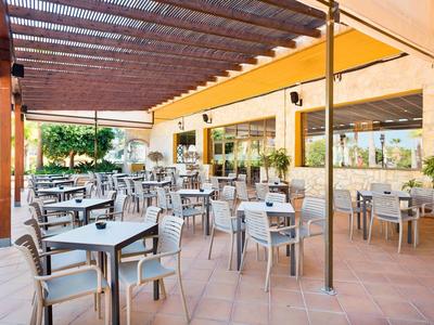 Freiluftrestaurant mit grauen Tischen und Stühlen, gelben Sonnensegeln und Terrakottaboden.